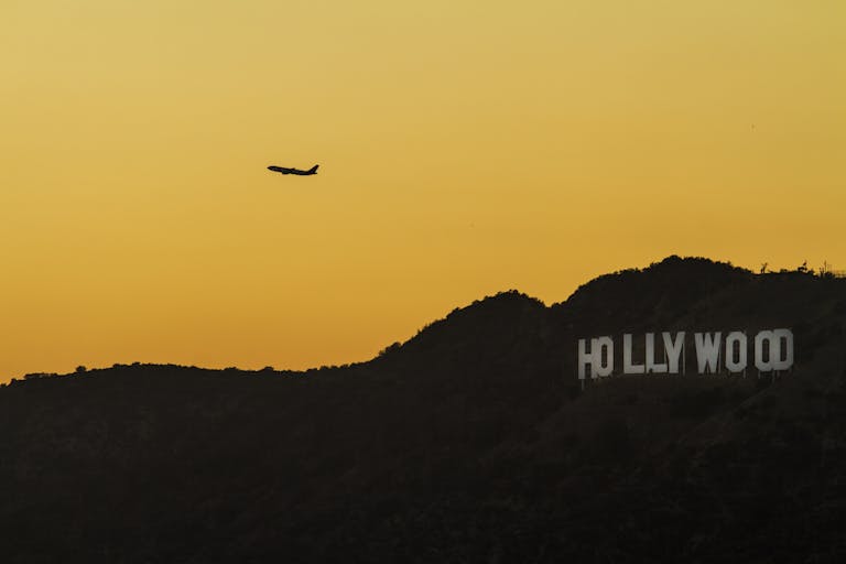 A silhouette of an airplane flying above the iconic Hollywood Sign against a vibrant sunset.